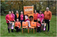 8 Frauen und ein Mann mit zwei orangefarbenen Stühlen und einem bunten Rollup. Alle tragen rosa und organgene Kleidung.